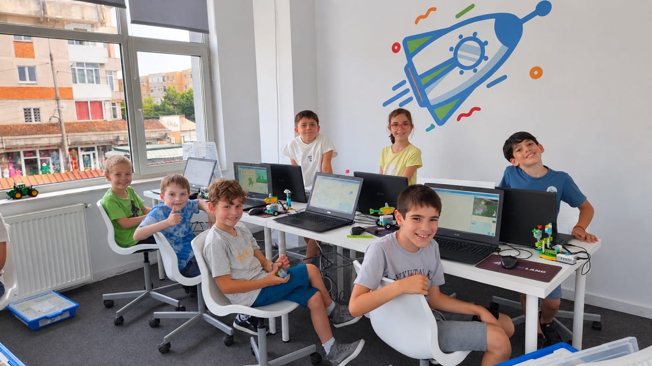 Copii zâmbitori la un curs Codeland, la laptopuri cu proiecte Scratch și roboți LEGO pe masă.