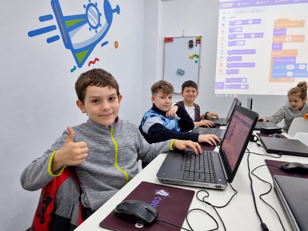 Copil la cursul de Programare Scratch Codeland, cu degetul mare ridicat, lângă colegii care lucrează la laptopuri.